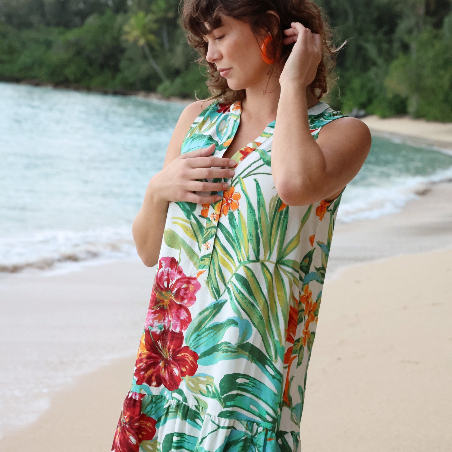 Mujer con vestido floral en una playa con el océano y vegetación al fondo.