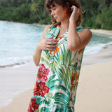 Mujer con vestido floral en una playa con el océano y vegetación al fondo.