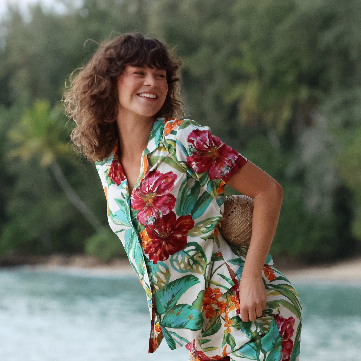 Mujer con un colorido vestido floral junto a un cuerpo de agua con vegetación al fondo.