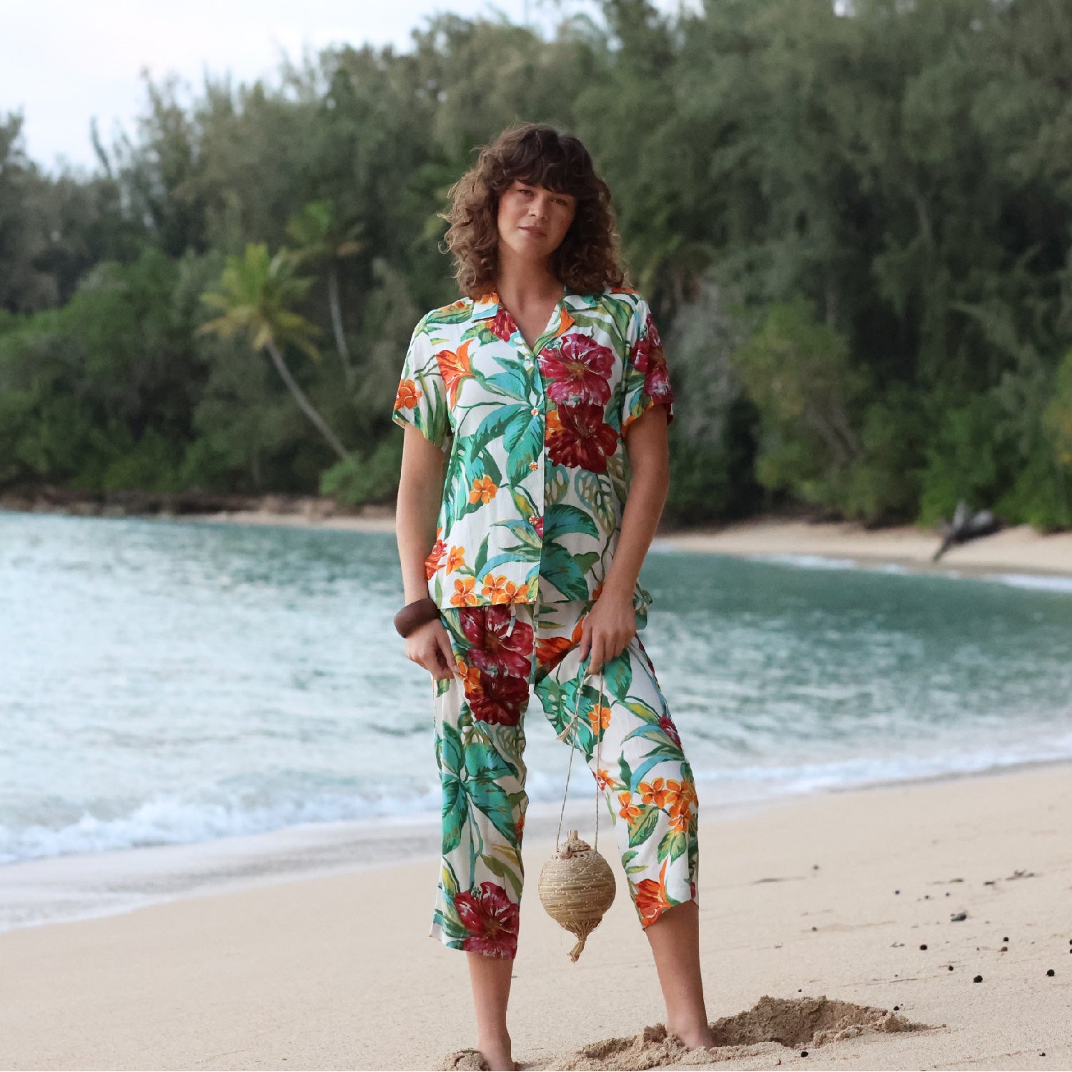 Woman in a colorful floral outfit standing on a beach with ocean and trees in the background
