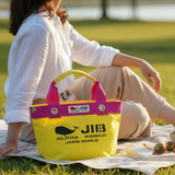 Un sac cabas jaune JIB à anses roses est posé sur une nappe de pique-nique blanche dans un parc herbeux. Derrière, une personne en chemise de lin blanc est assise, des lunettes de soleil et un livre à portée de main, baignée par la douce lumière de l'après-midi.