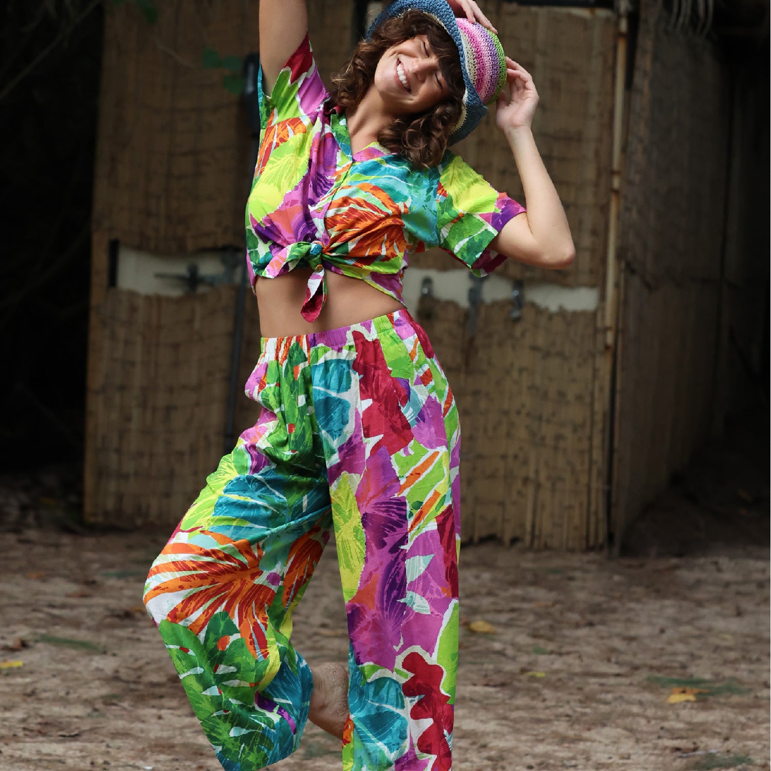 Person wearing a colorful tie-dye outfit with a hat, standing in front of a wooden wall.