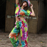 Person wearing a colorful tie-dye outfit with a hat, standing in front of a wooden wall.