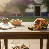 Colorful cap on a wooden table with a cup, book, and bread basket outdoors.