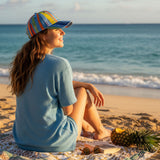 Woman sitting on a beach with a colorful cap and pineapple, looking at the ocean.