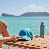 Person sitting by a table with a cap, sunglasses, and water bottle on a beach with mountains in the background