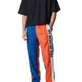 Persona que viste una camiseta negra y pantalones deportivos azules y naranjas con un logotipo de marca visible sobre un fondo blanco.