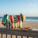 Un bolso de rayas de colores, un sombrero de paja y unas sandalias sobre un banco de madera en la playa.
