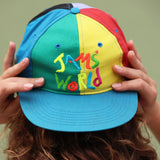 Gorra colorida con el texto 'JAM'S WORLD' sostenida por una persona sobre un fondo liso.