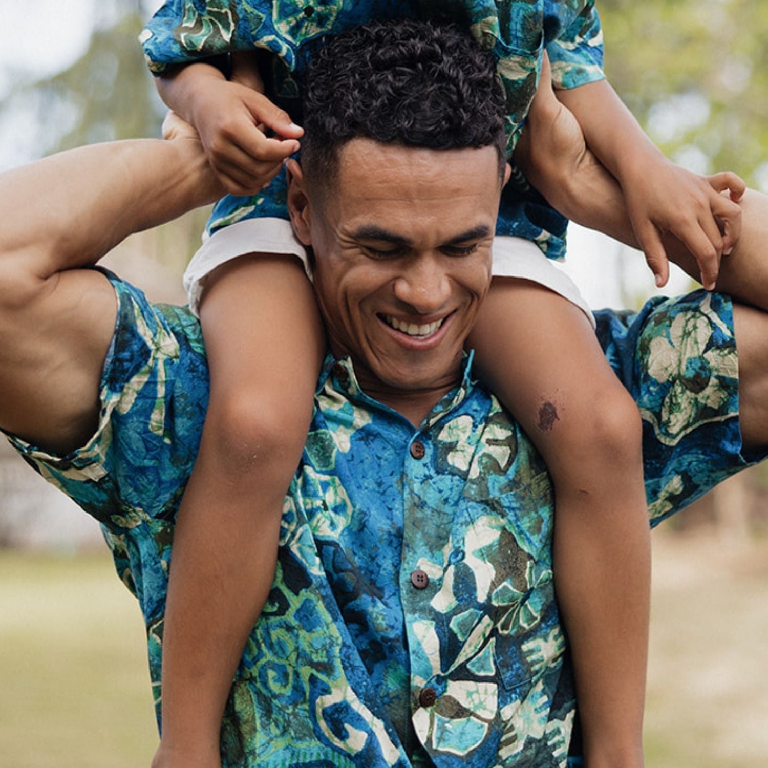 Man carrying a child on his shoulders outdoors, both wearing matching blue floral outfits.