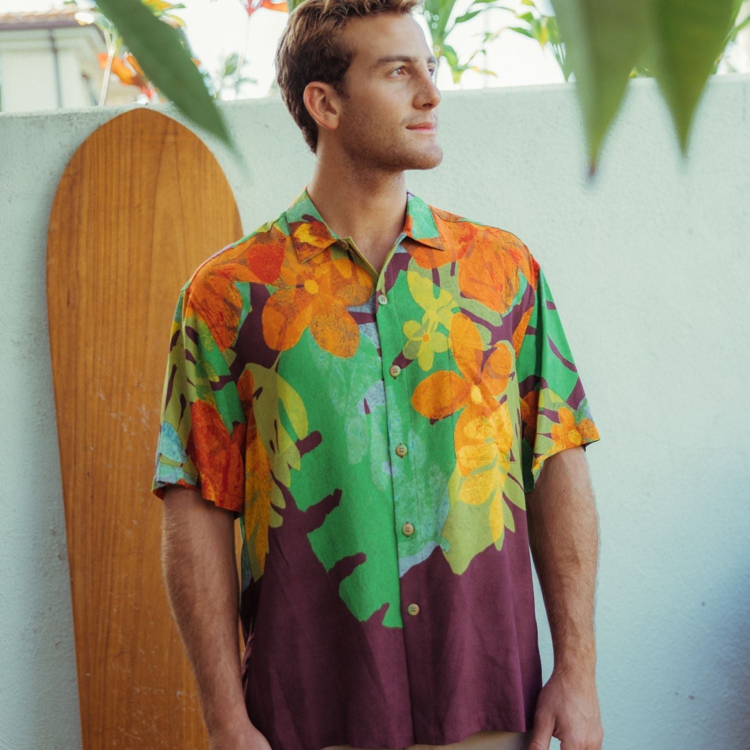 Hombre vestido con una camisa floral colorida de pie frente a una pared de color claro con una tabla de surf de madera.