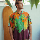 Hombre vestido con una camisa floral colorida de pie frente a una pared de color claro con una tabla de surf de madera.