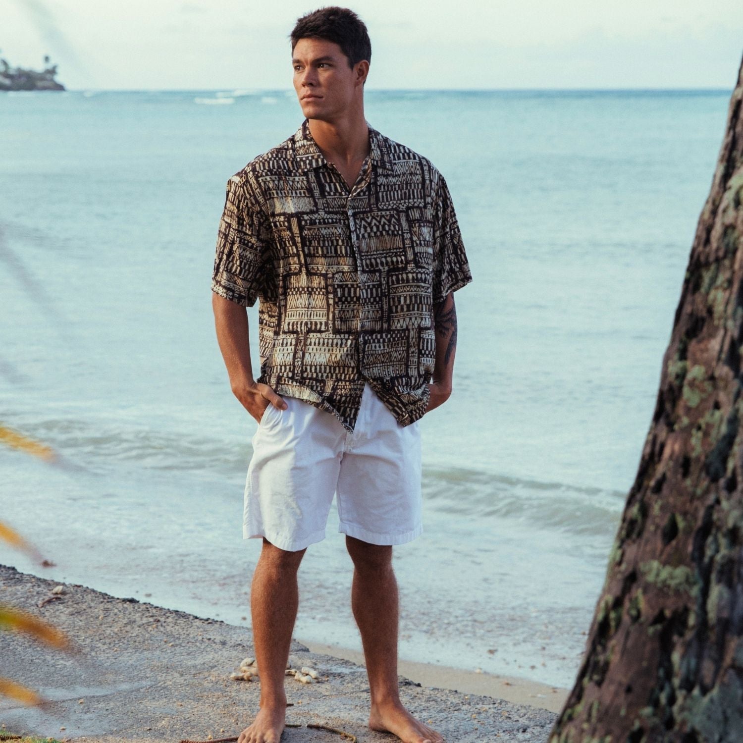 Hombre con camisa estampada y pantalones cortos blancos de pie en una playa.