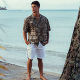 Hombre con camisa estampada y pantalones cortos blancos de pie en una playa.