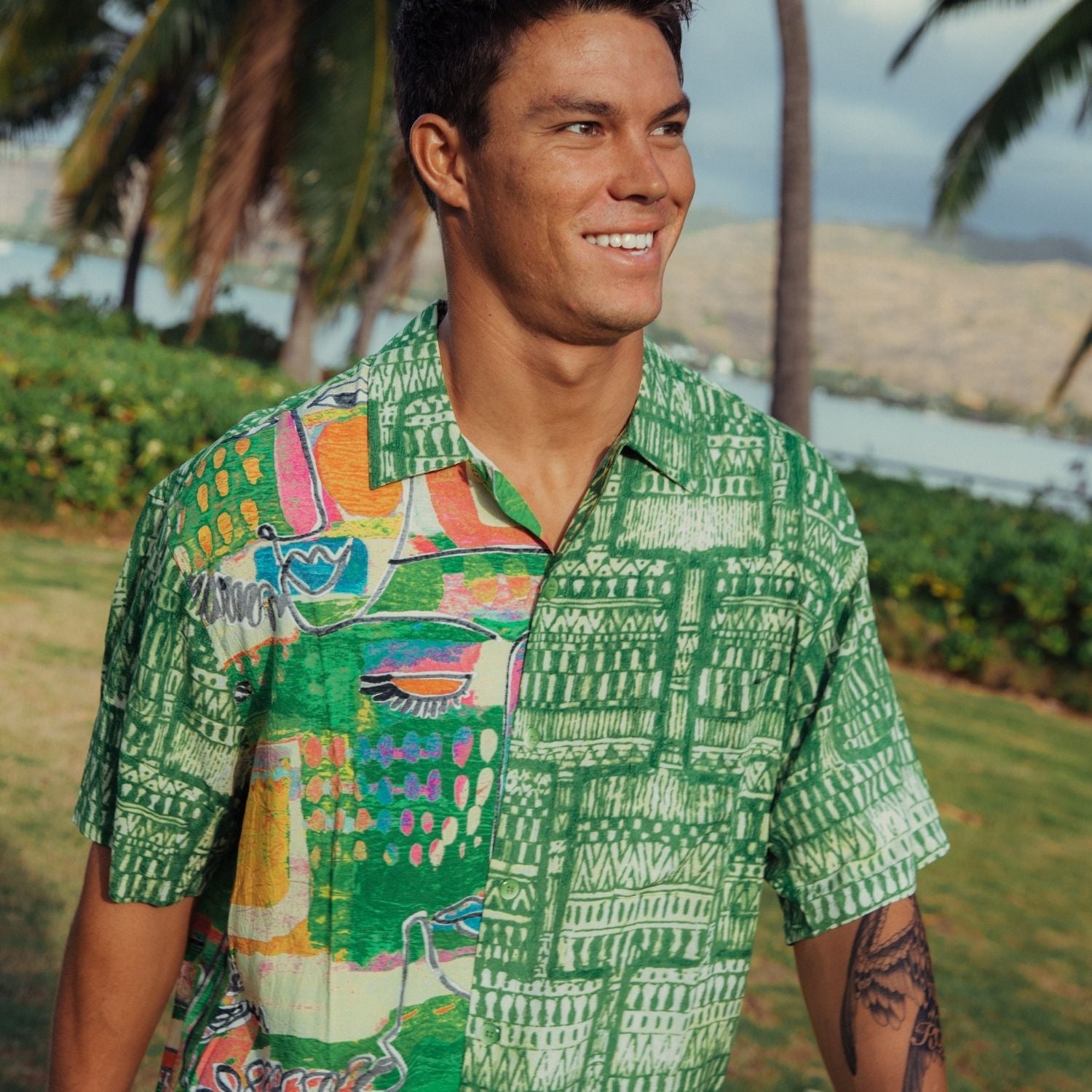 Man wearing a colorful patterned shirt in a tropical setting with palm trees and water.
