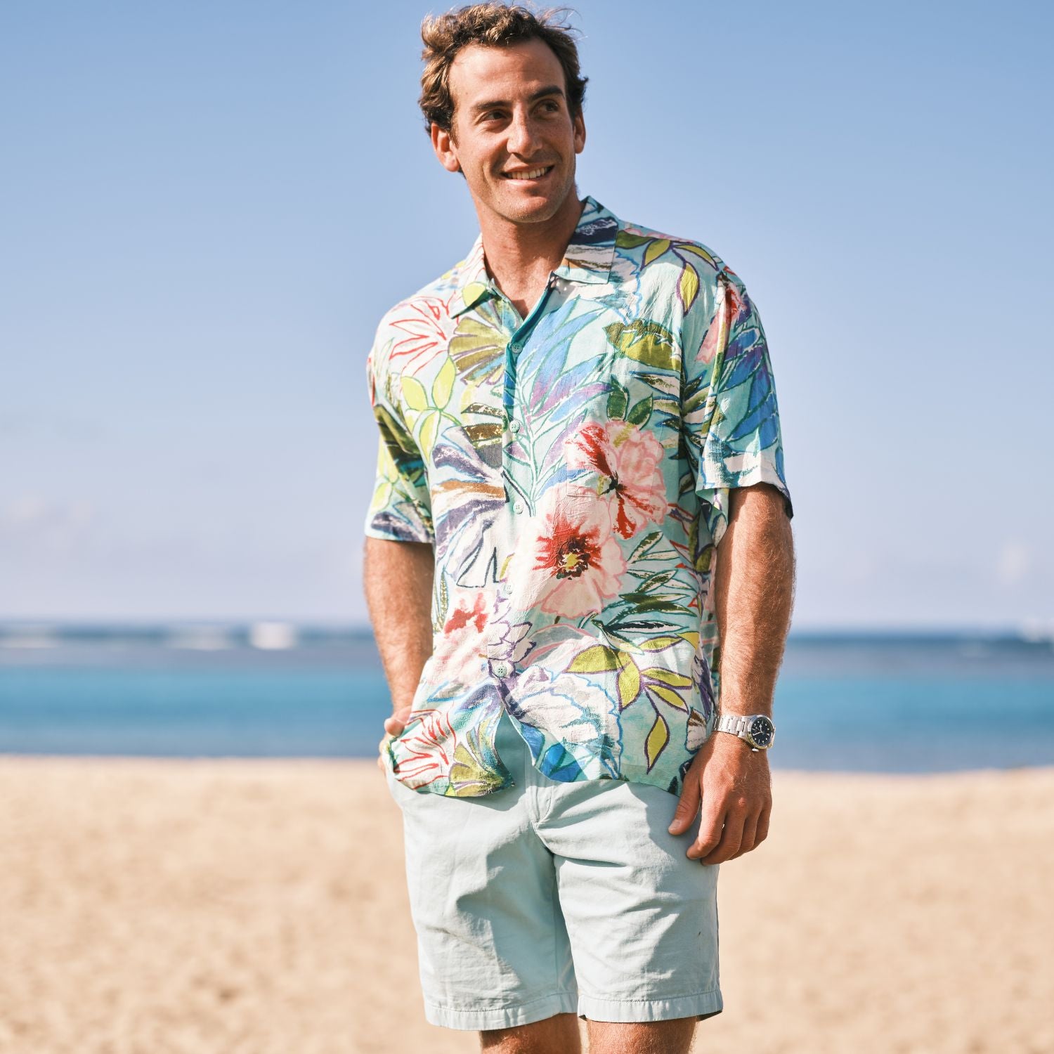 Man wearing a colorful floral shirt and light shorts standing on a beach.