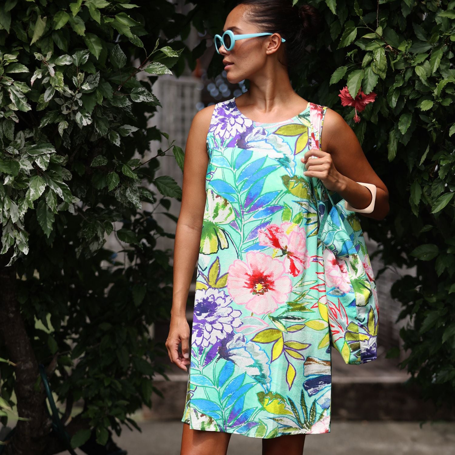 Woman wearing a colorful floral dress and sunglasses standing among greenery.