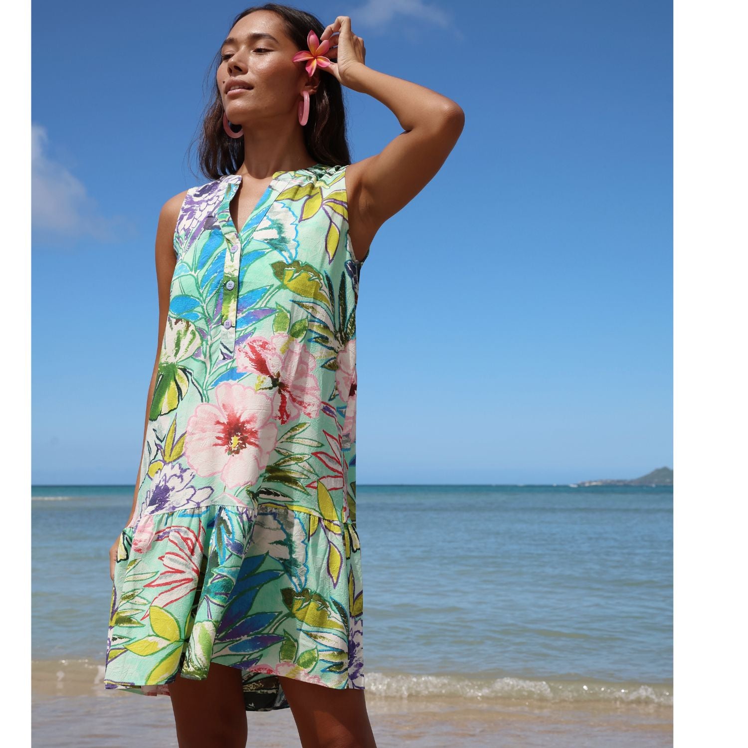 Woman in a colorful floral dress standing on a beach with clear blue sky and ocean.