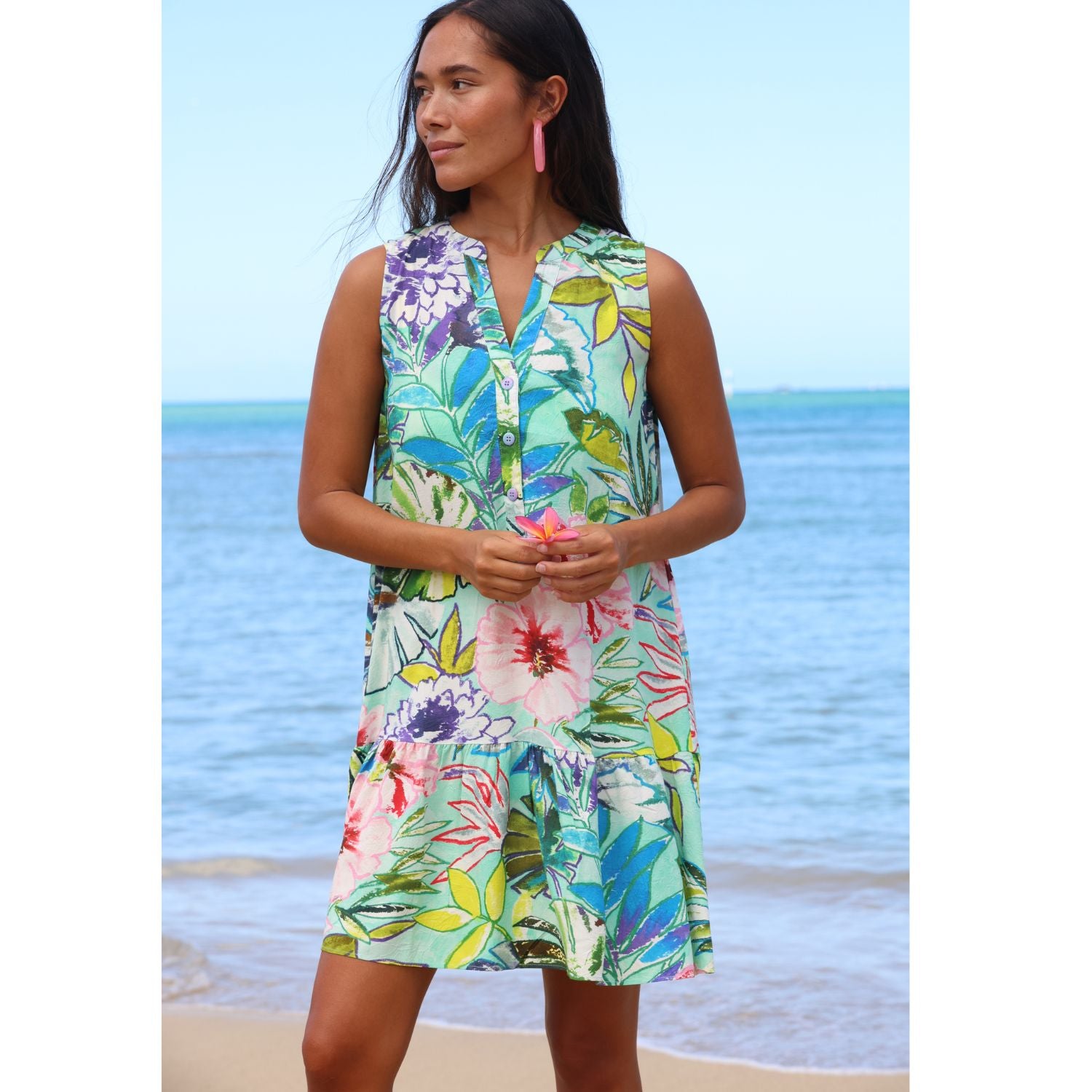 Woman wearing a colorful floral dress standing on a beach with blue water and sky.
