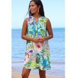 Woman wearing a colorful floral dress standing on a beach with blue water and sky.