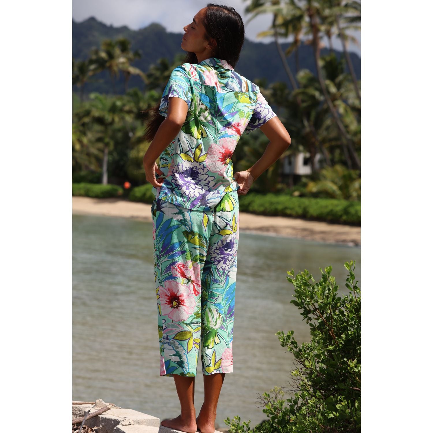 Woman in a colorful floral jumpsuit standing by a body of water with palm trees in the background.