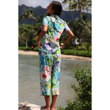 Woman in a colorful floral jumpsuit standing by a body of water with palm trees in the background.
