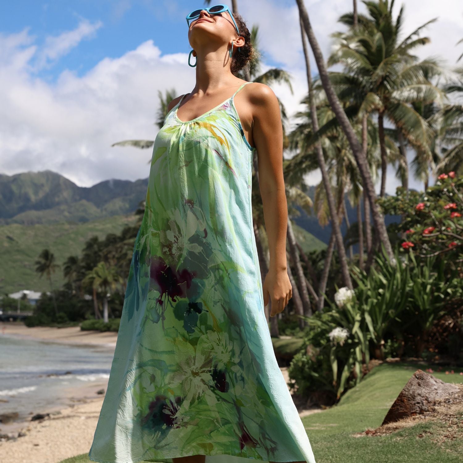 Woman in a floral dress standing on a beach with palm trees and mountains in the background