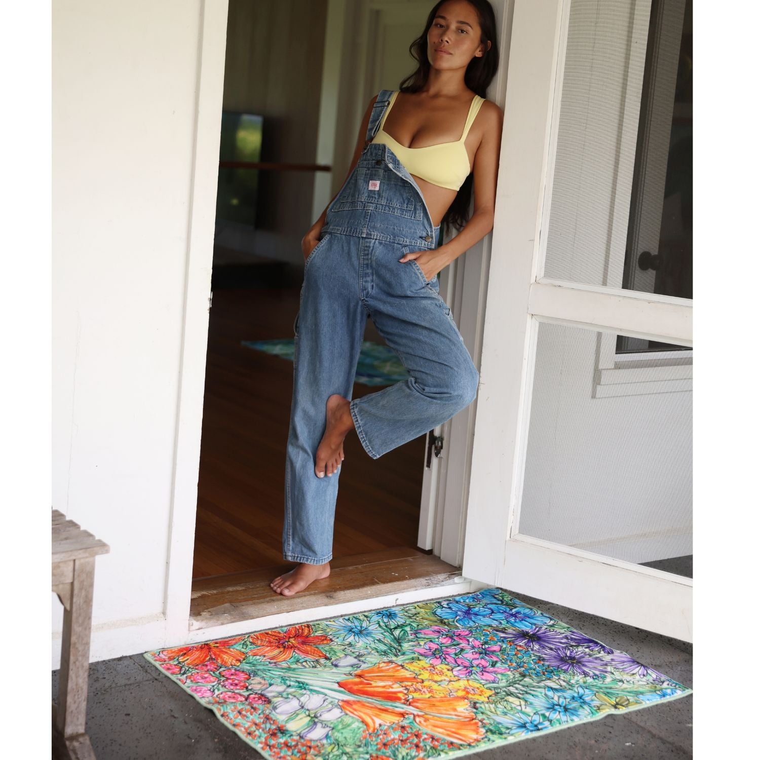 Woman in denim overalls and yellow top standing on a colorful doormat.