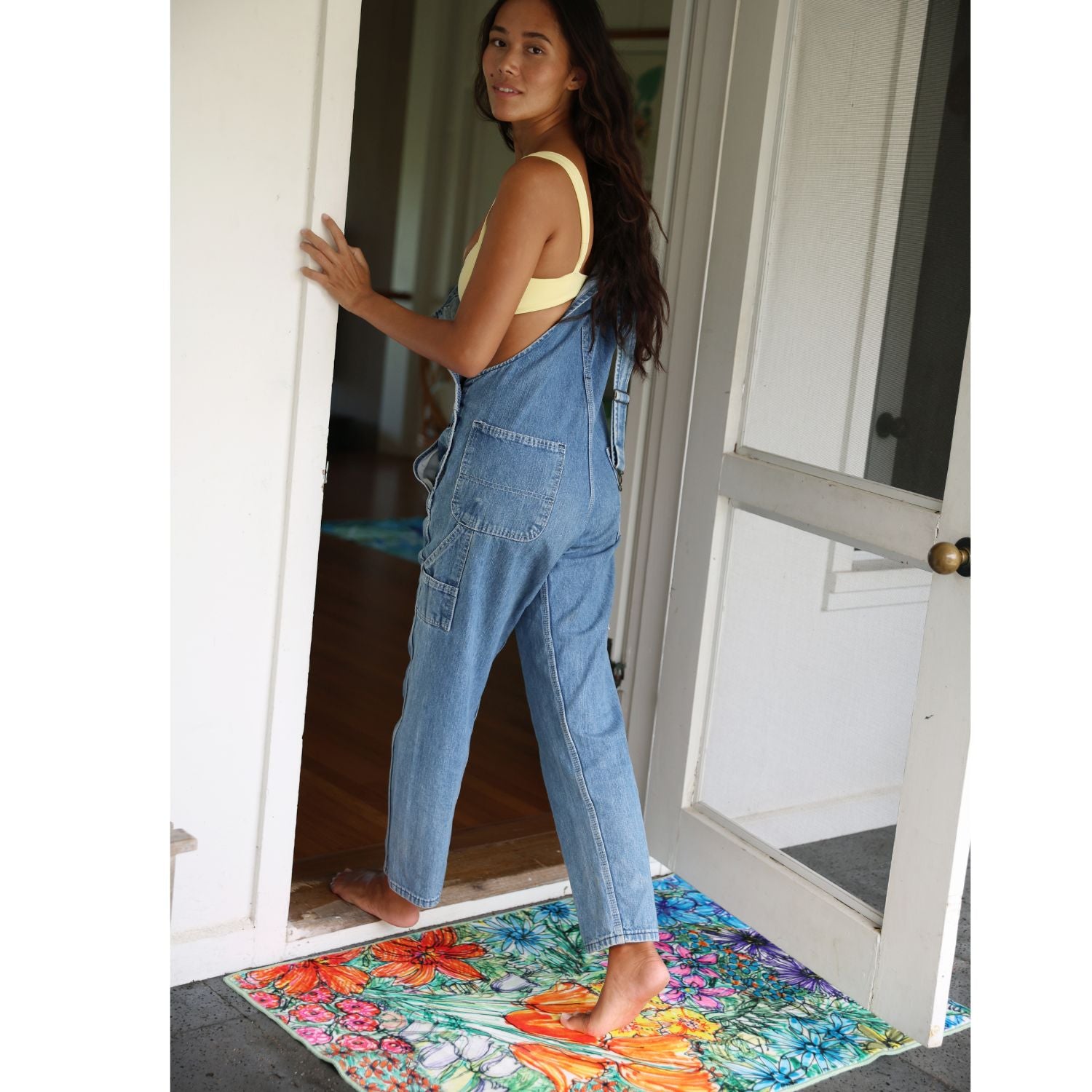 Woman in blue overalls stepping onto a colorful doormat from a doorway.