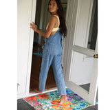 Woman in blue overalls stepping onto a colorful doormat from a doorway.