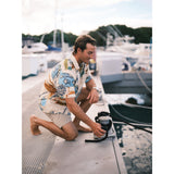 Man in a colorful shirt and shorts kneeling on a dock with boats in the background