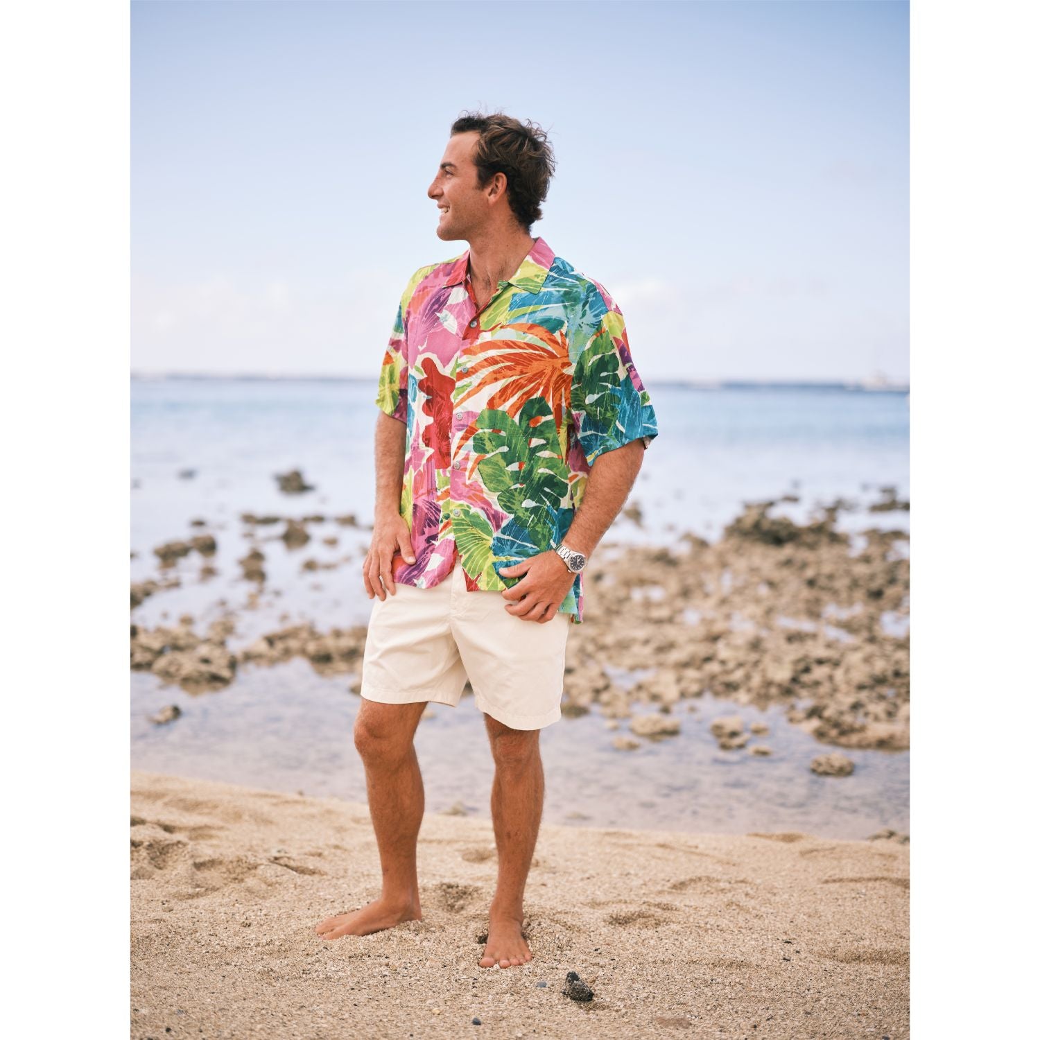 Hombre con una colorida camisa tropical y pantalones cortos blancos de pie en una playa.