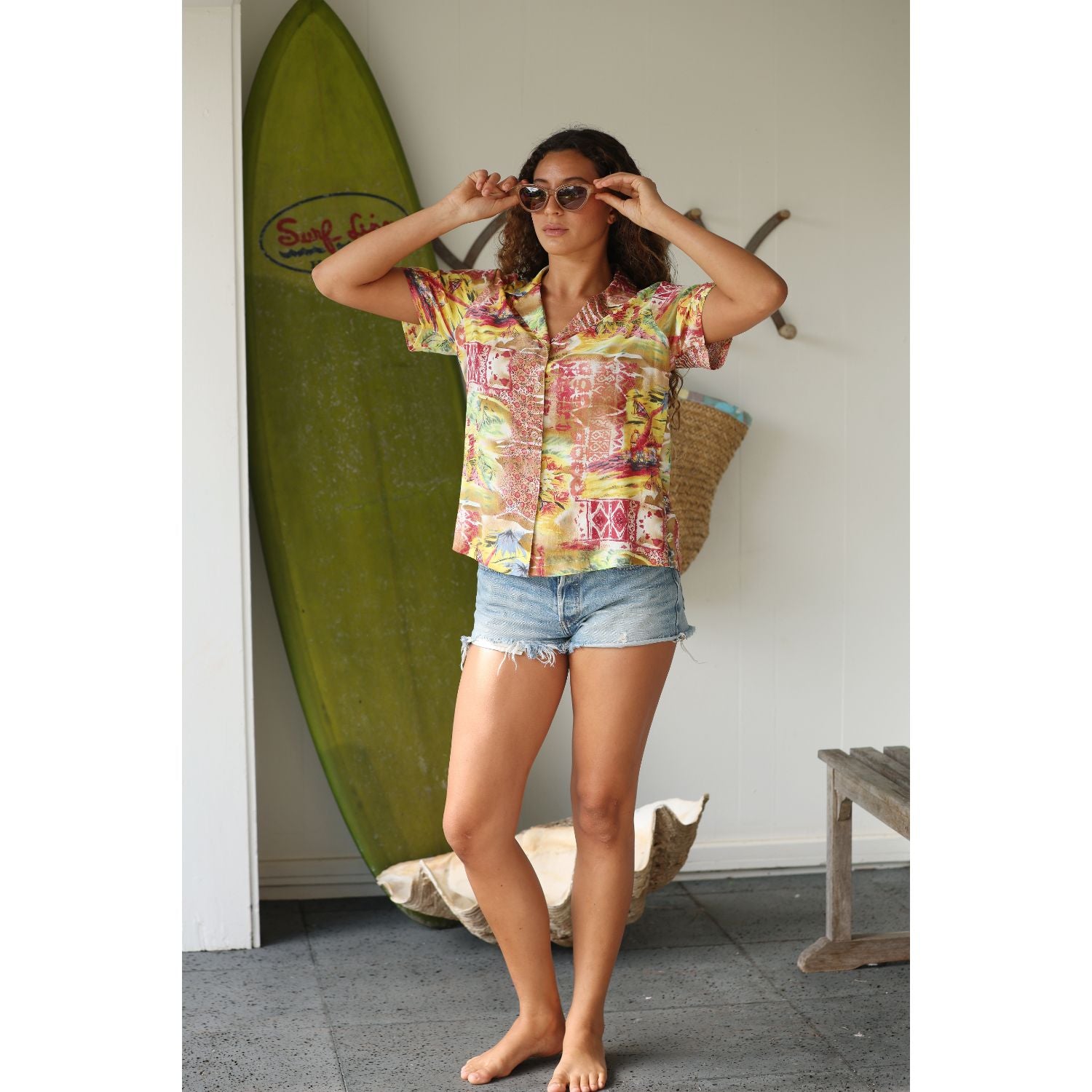 Woman in a colorful shirt and denim shorts standing next to a green surfboard indoors.