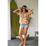 Woman in a colorful shirt and denim shorts standing next to a green surfboard indoors.
