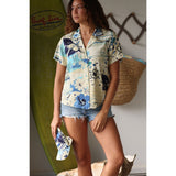Short-sleeve shirt with blue and white floral pattern on a beige background on model with jeans shorts matching cap  standing infront of surf board and shell.