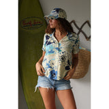 Short-sleeve shirt with blue and white floral pattern on a beige background on model with jeans shorts matching cap  standing infront of surf board.