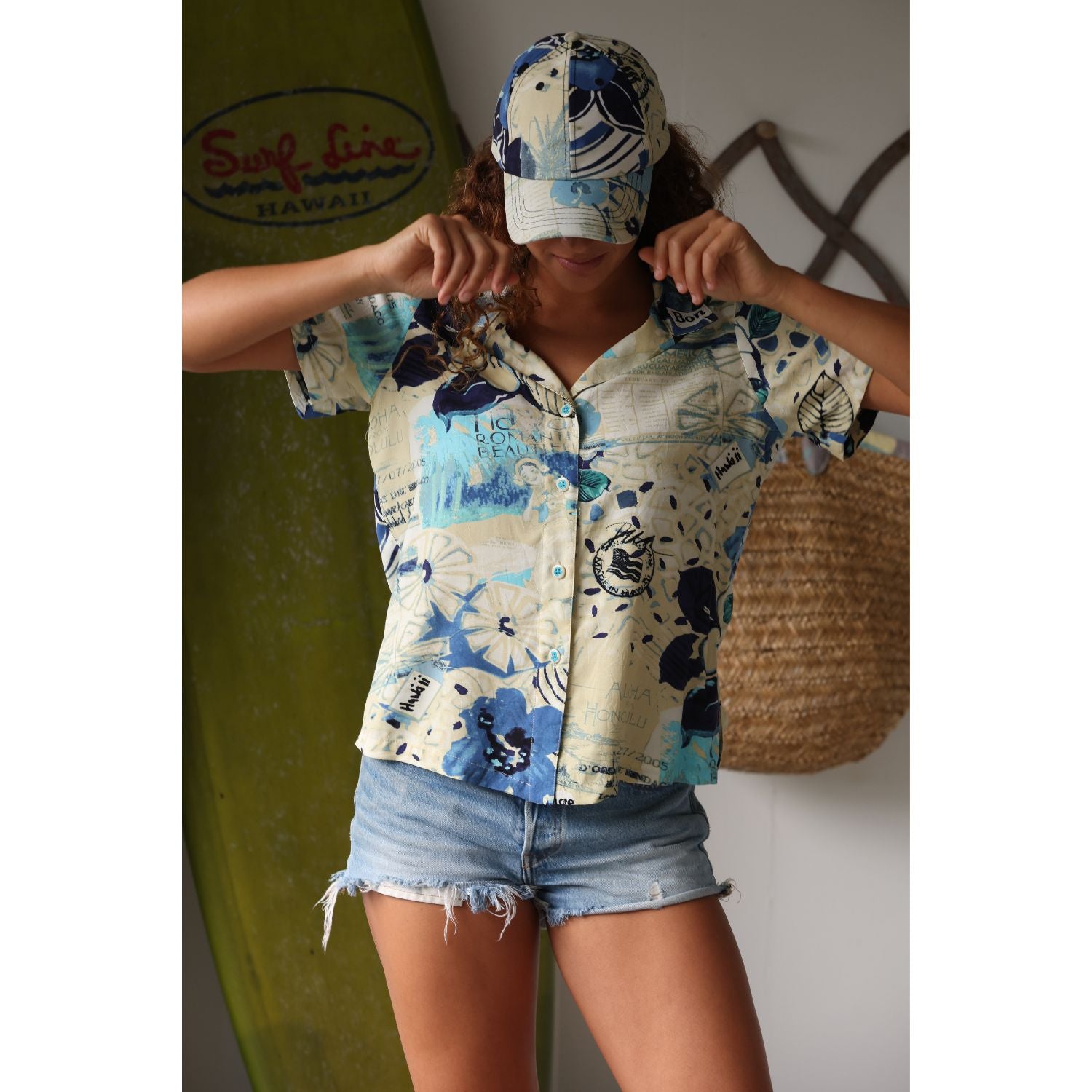 Short-sleeve shirt with blue and white floral pattern on a beige background on model with jeans shorts matching cap  standing infront of surf board and shell.