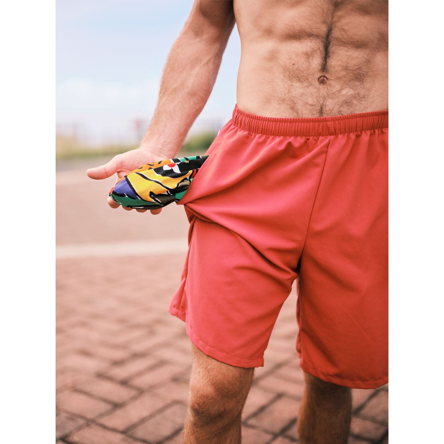 Model standing on walk way with solid red shorts with his shorts pocket pulled out which is in a colorful print