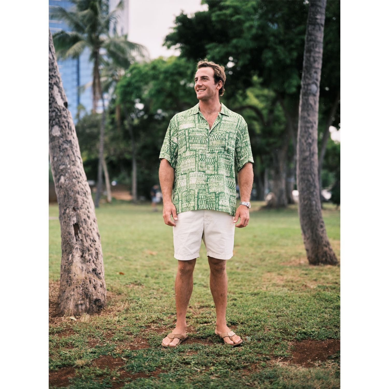Man wearing a green patterned shirt and white shorts standing in a park with trees and buildings in the background.