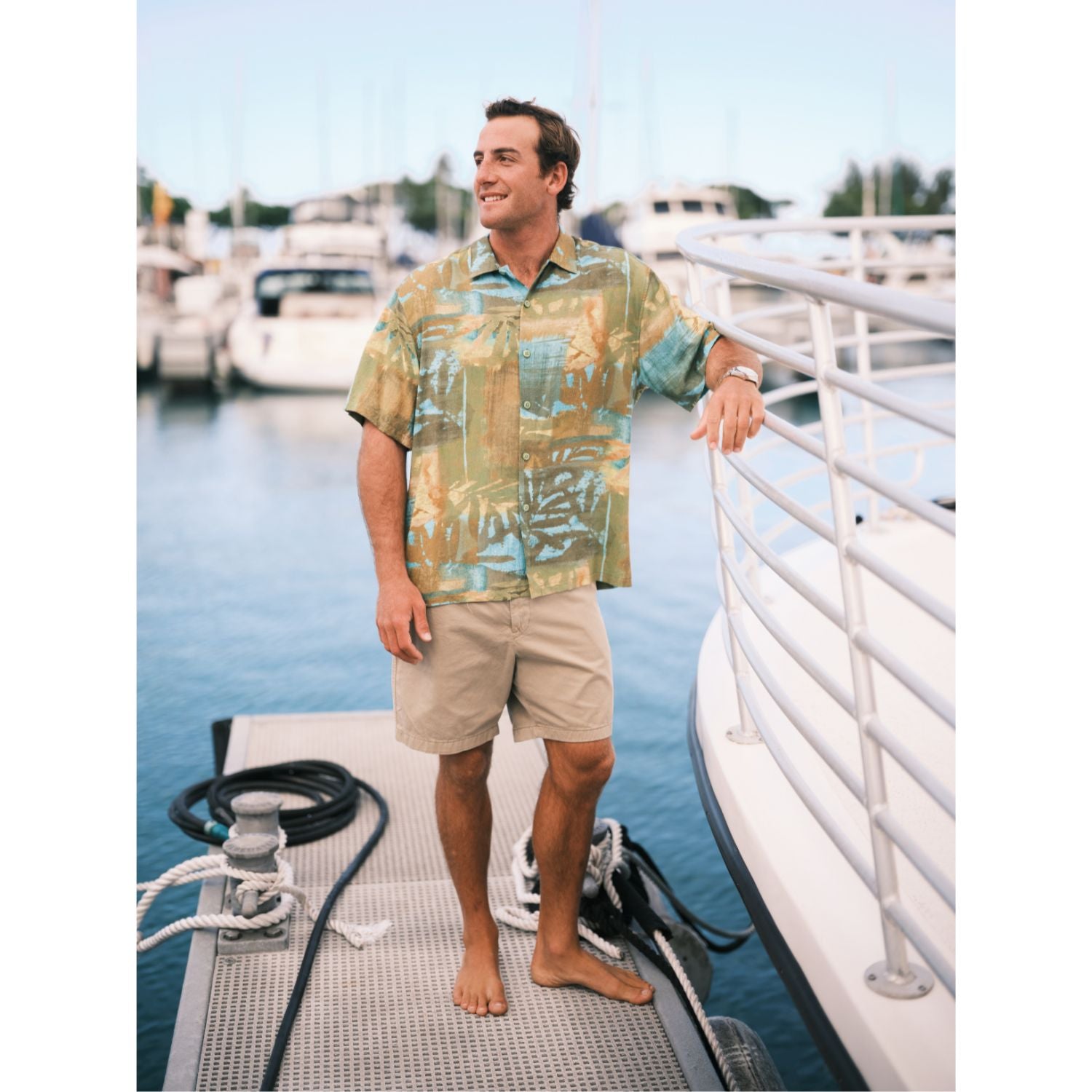 Model wearing Fern Patch button down Aloha shirt, an Abstract art with teal, orange, and green brush strokes. solid Khaki shorts standing on pier.