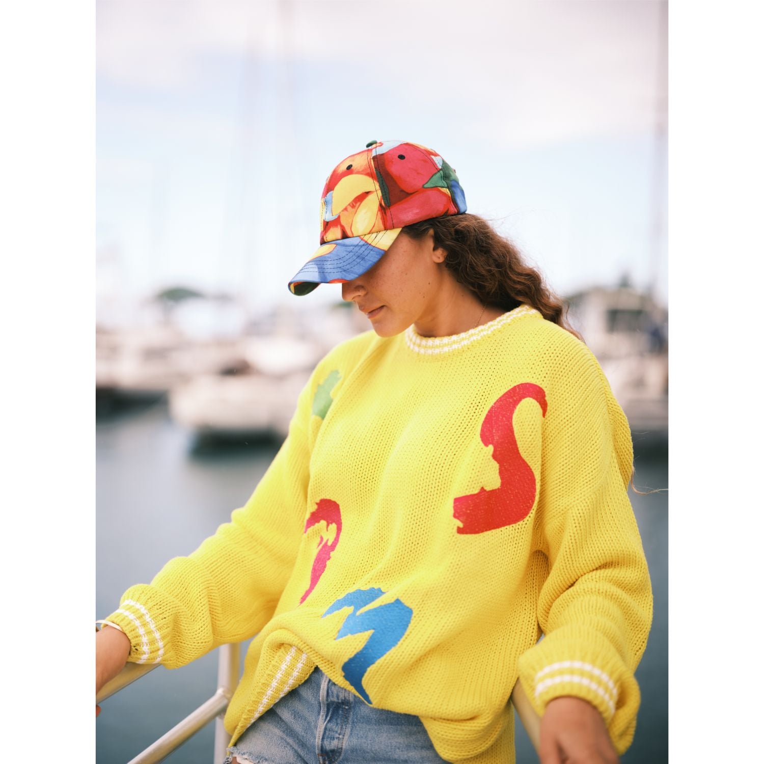 Person wearing a yellow sweater with colorful letters and a multicolored cap by a waterfront.