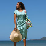 Woman in a light blue dress holding a straw hat and a colorful bag on a beach with clear blue sky and ocean.