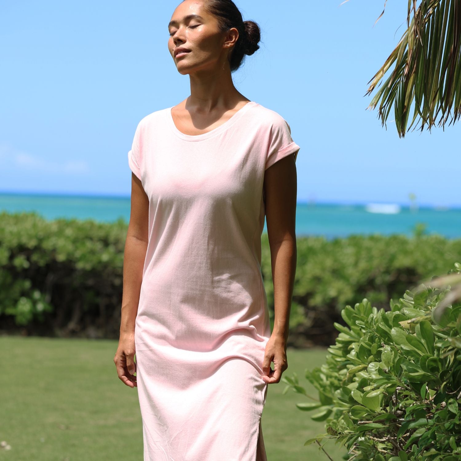 Woman in a pink dress standing outdoors with greenery and blue sky in the background