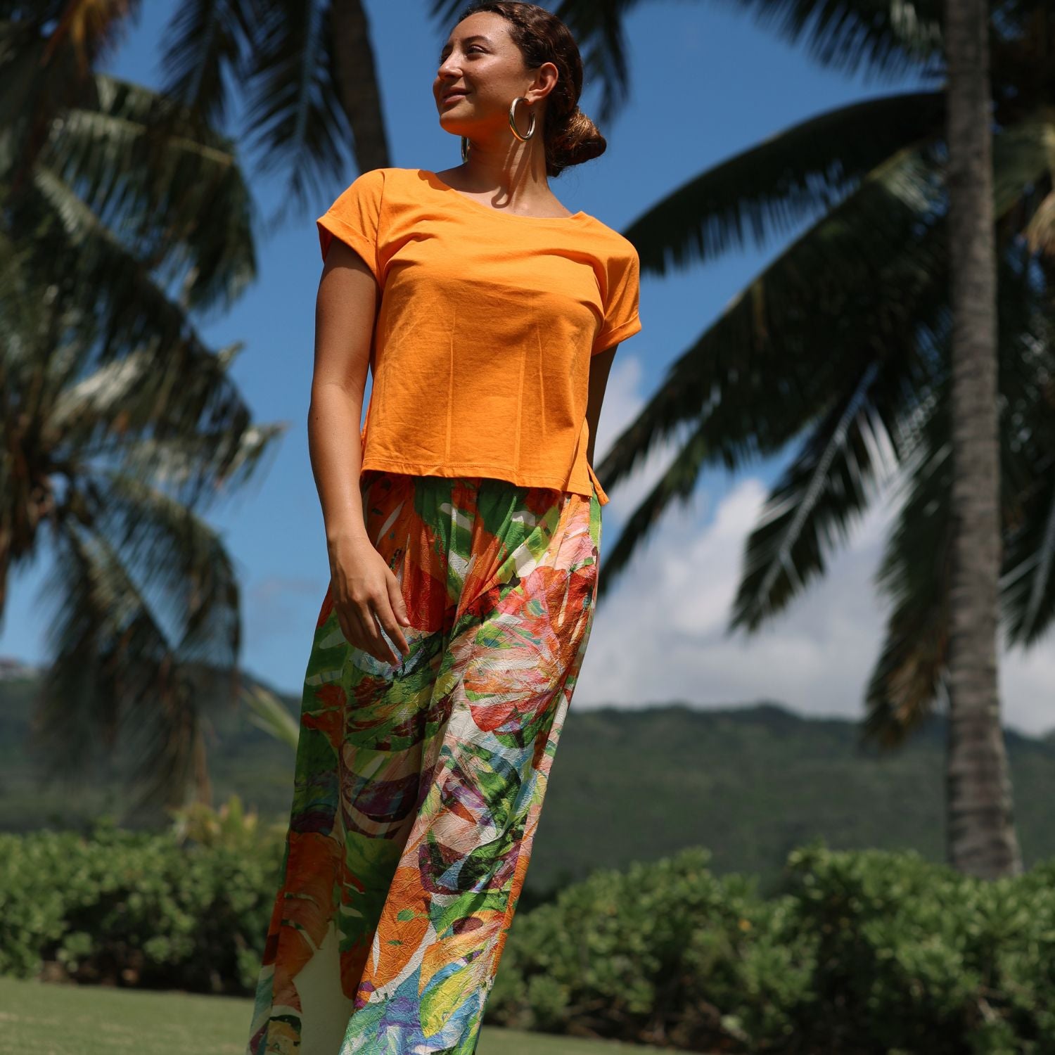 Woman wearing an orange top and colorful floral pants standing outdoors with palm trees and mountains in the background.