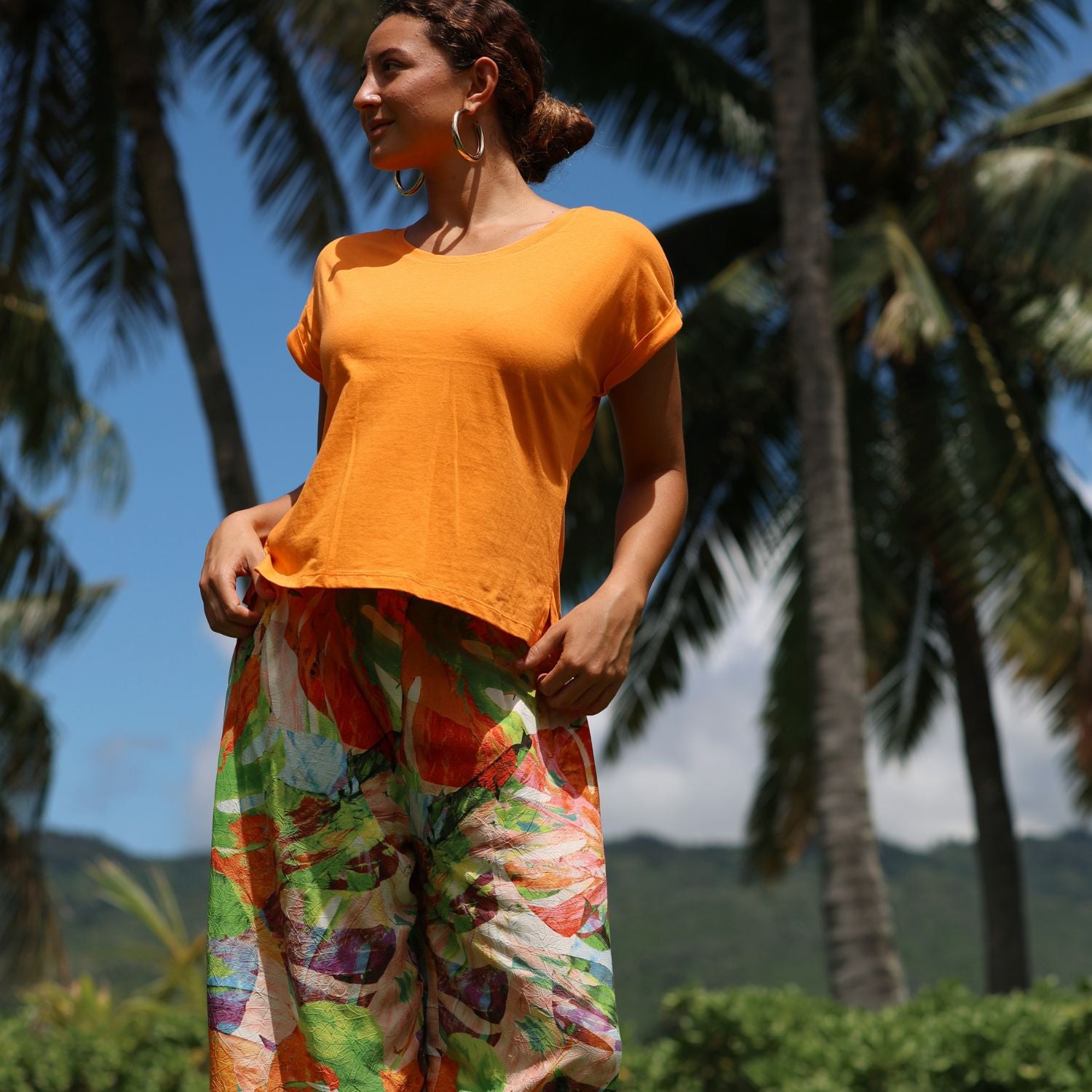 Woman in an orange top and colorful pants standing outdoors with palm trees in the background