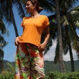 Woman in an orange top and colorful pants standing outdoors with palm trees in the background