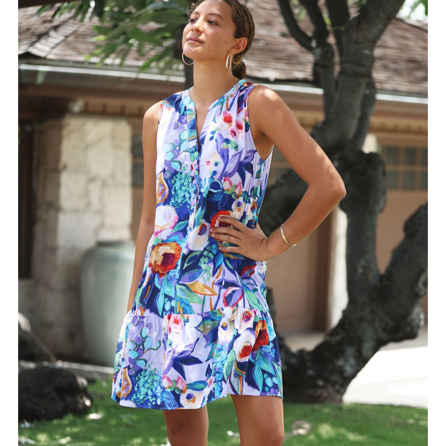 A woman wearing a sleeveless, knee-length floral dress with a vibrant multicolor print of blue, purple, red, and white flowers, standing outdoors in front of a house and a tree.