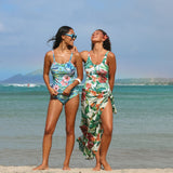 Two women standing on a sandy beach wearing tropical floral swimsuits and a matching floral dress, with the ocean and distant land visible in the background.