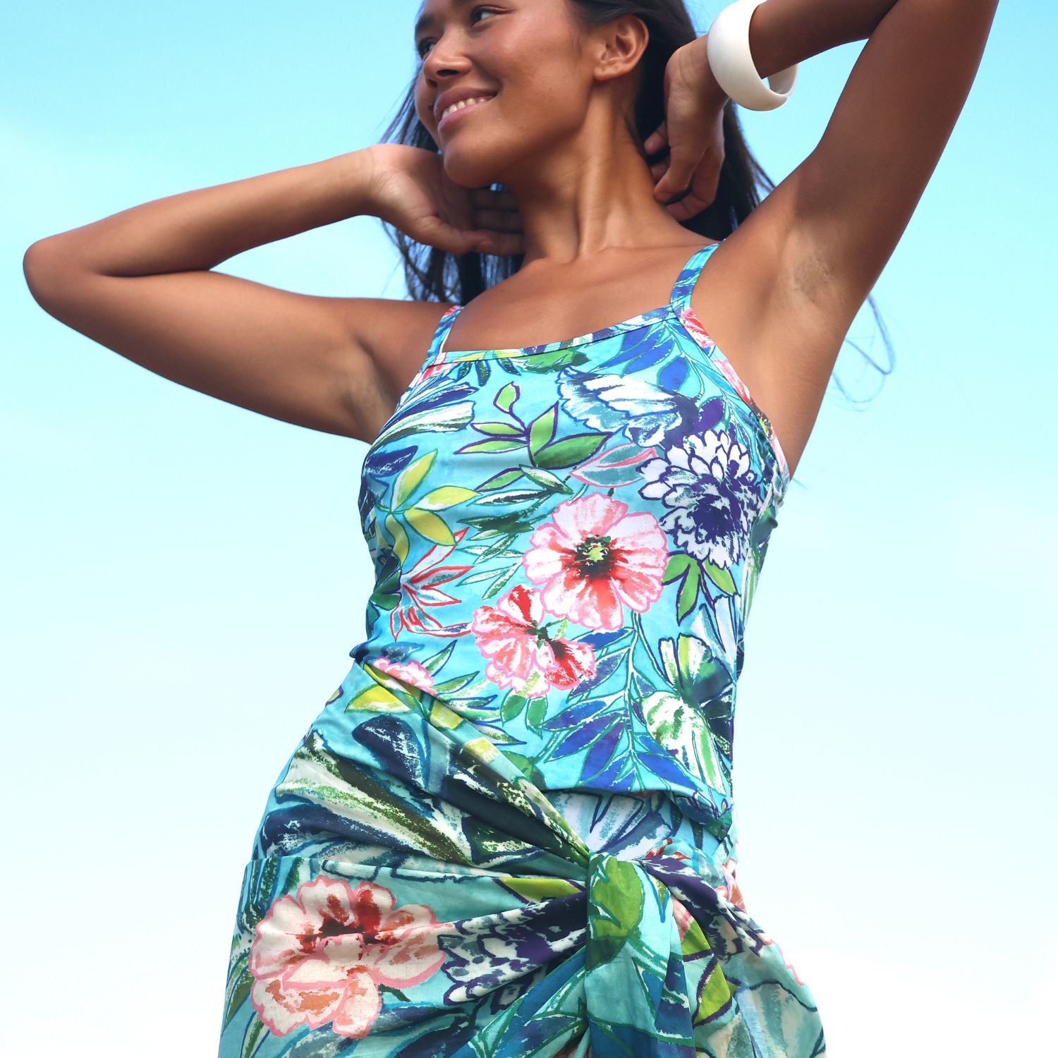 A woman wearing a sleeveless tropical floral print dress with a gathered waist, set against a bright light blue sky background.