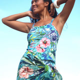 A woman wearing a sleeveless tropical floral print dress with a gathered waist, set against a bright light blue sky background.